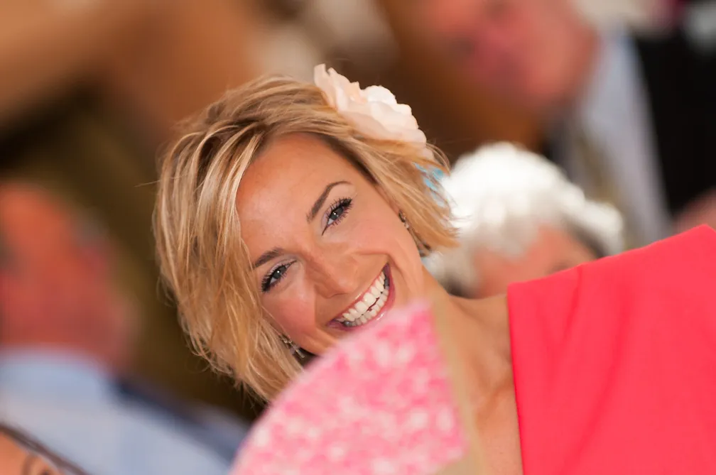 Smiling woman with short blonde hair and a white flower hair accessory, holding a pink fan.