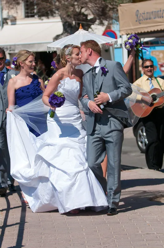 Bride in white wedding dress kissing groom in gray suit while bridesmaids and a guitarist celebrate outdoors.