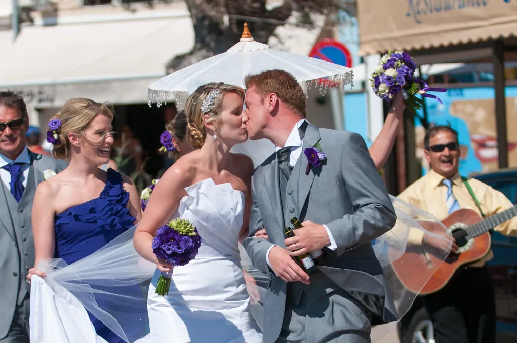 Bride and groom kissing outdoors with bridal party and a man playing guitar in the background.