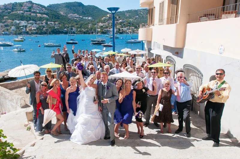 Large group of wedding guests, including bride in white dress and groom in gray suit, posing on stone steps by the water with boats and hills in the background.