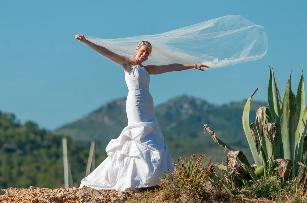 Bride in a strapless white wedding dress standing on rocky ground holding her veil spread wide with mountains and clear sky in the background.