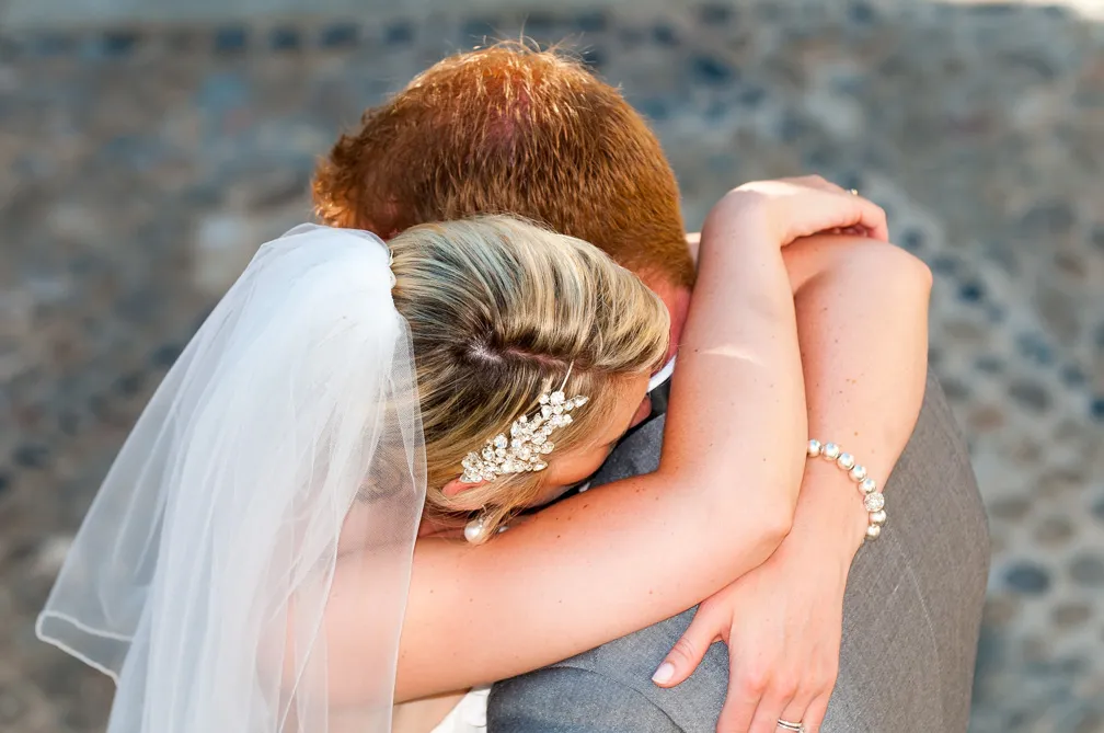 Bride with a veil and pearl bracelet embracing groom with red hair in a gray suit.