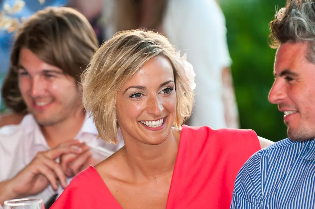 Smiling woman with short blonde hair wearing a red top, seated between two men at a social event.