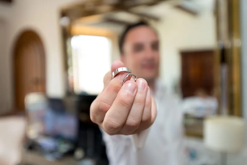 Close-up of a hand holding two wedding rings in a living room with a blurred person in the background.