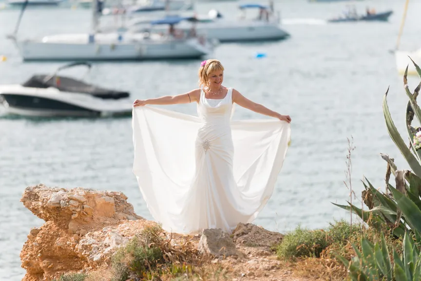 Woman in a white wedding dress standing on rocky ground by the water, holding out the sides of her dress with boats in the background.