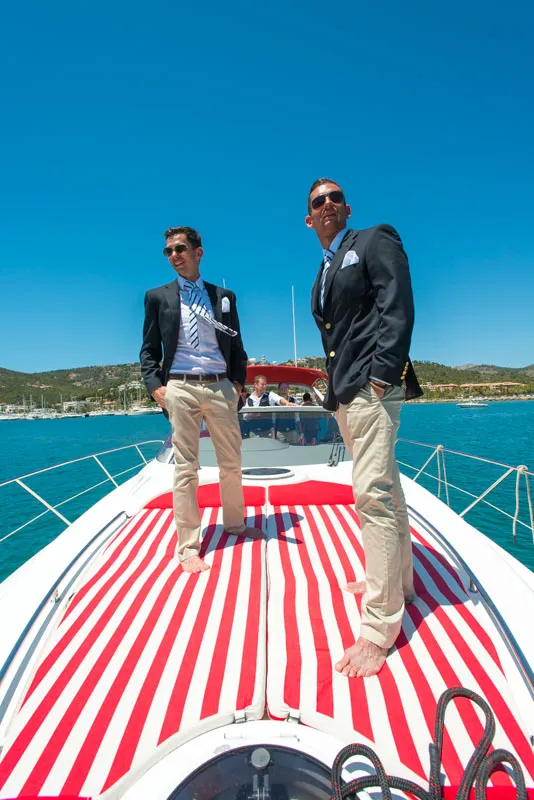 Two men in blazers and beige pants standing barefoot on a red-and-white striped mat on a yacht under clear blue sky.