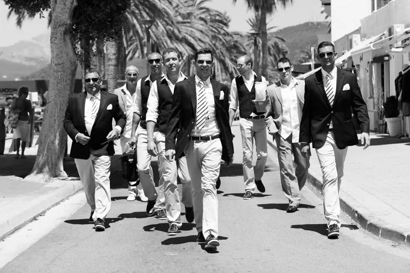 Seven men dressed in suits and casual formal wear walking confidently down a sunny street lined with palm trees.
