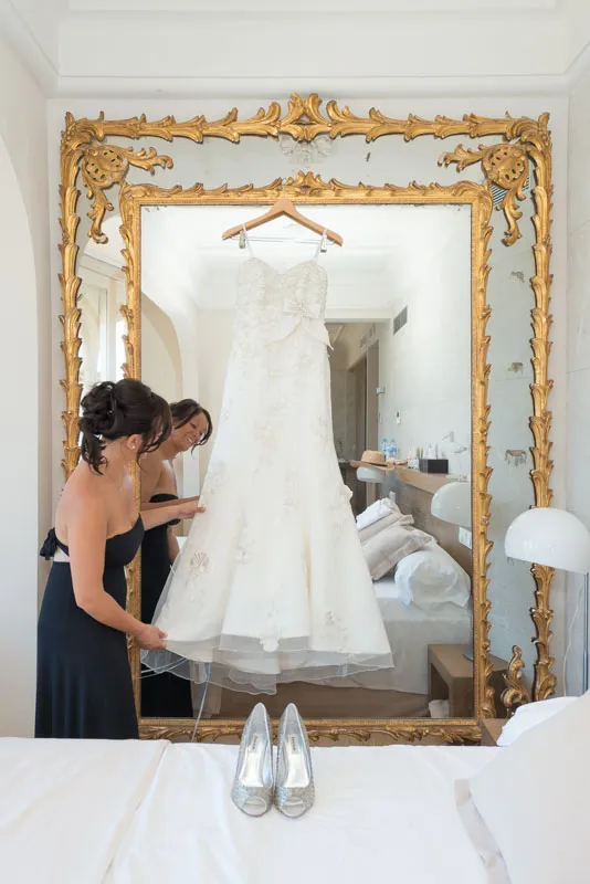 Bride in a black dress holding the hem of a white wedding gown hanging on an ornate gold mirror.