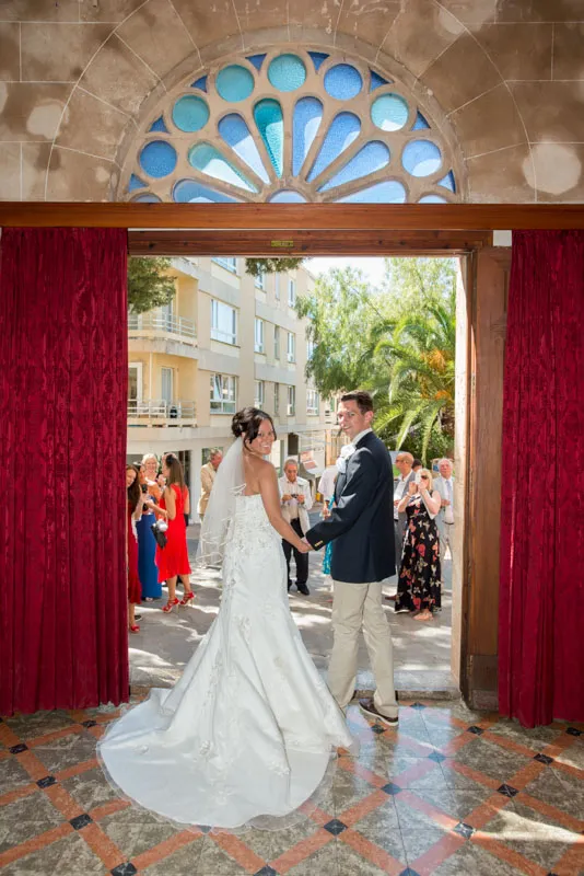Bride in white wedding gown and groom holding hands, standing in an open doorway with red curtains, facing guests outside.