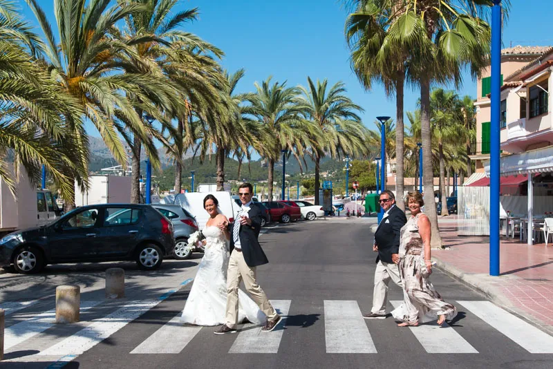 Bride and groom crossing a zebra crossing on a sunny street lined with palm trees, accompanied by an older couple.