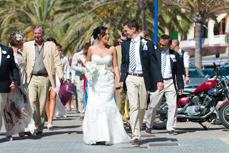 Bride and groom smiling and holding hands while walking with guests near palm trees on a sunny day.