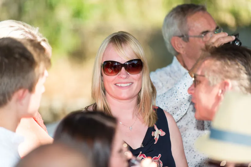Smiling woman wearing large sunglasses and a floral top surrounded by a group of people in an outdoor setting.