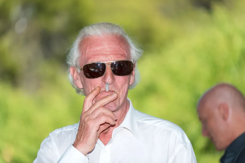 Older man with white hair and sunglasses smoking a cigarette outdoors with blurred person in background.