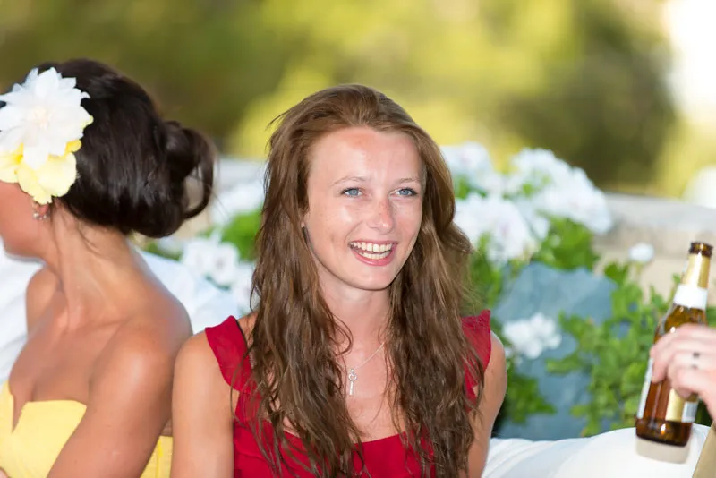 Smiling woman with long brown hair wearing a red dress, sitting outdoors with blurred greenery and white flowers in the background.