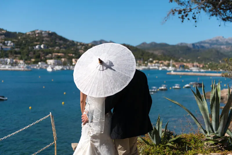 Couple embracing with the woman holding a white parasol, overlooking a scenic harbor with boats and distant hills.