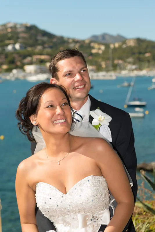 Smiling bride in a strapless white wedding dress and groom in a dark suit with a white flower boutonniere posing by a waterfront with boats and hills in the background.