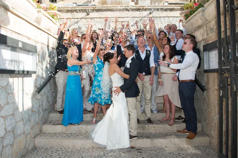 Bride and groom embrace on stone steps surrounded by joyful guests throwing flower petals at an outdoor wedding.