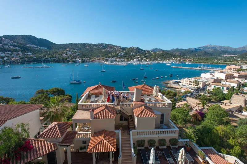 A coastal villa with terracotta roofs overlooking a bay filled with anchored sailboats and surrounded by green hills under a clear blue sky.