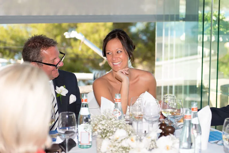 Smiling bride sitting at a table with a man wearing glasses and a suit, surrounded by glassware and floral decorations.