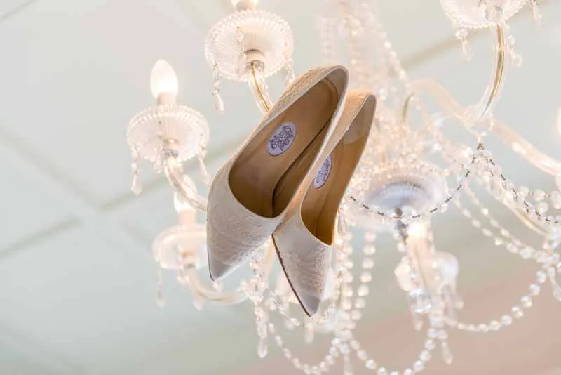Pair of elegant white lace bridal shoes hanging on a crystal chandelier.