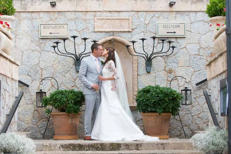 Bride in white dress and groom in light gray suit kissing on stone steps between potted plants under signs pointing to restaurant and reception.