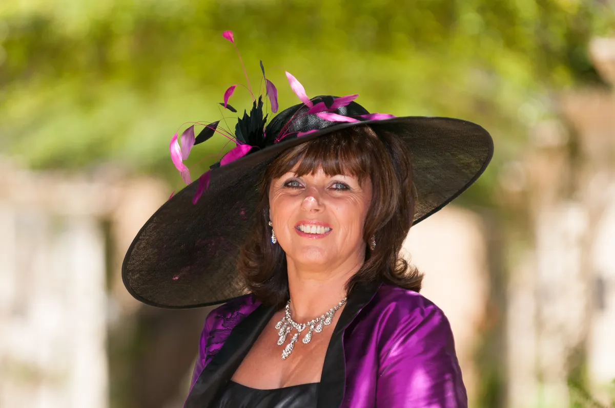 Woman wearing a large black hat adorned with pink feathers and a purple satin jacket, smiling outdoors.