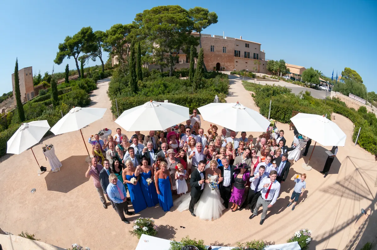 Large wedding group with bride and groom in center, guests dressed formally, standing outdoors under white umbrellas on sunny day.