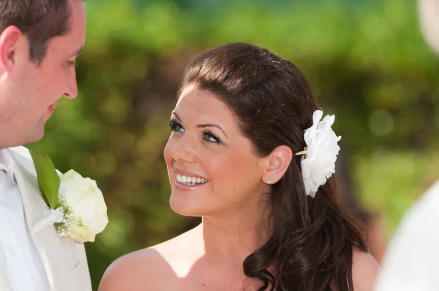 Bride with dark hair and white flower in her hair smiling and looking at groom wearing a white suit with a white rose boutonniere.