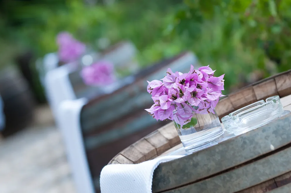 Purple bougainvillea flowers in a glass vase on a wooden barrel with a white cloth and glass ashtray.