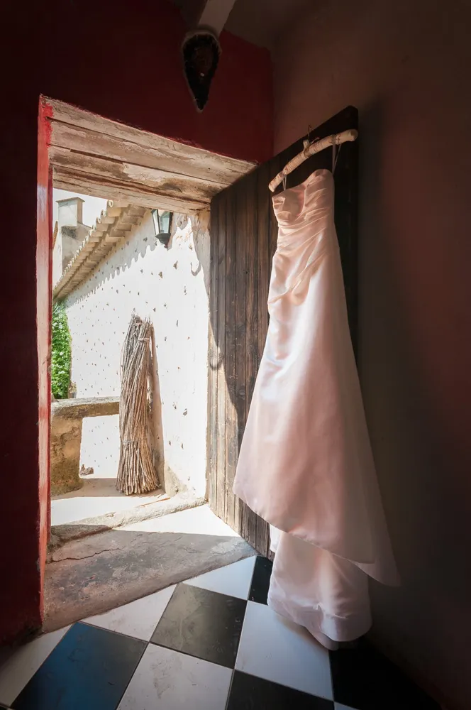 White strapless wedding dress hanging on a wooden door frame in a rustic room with black and white checkered floor tiles.