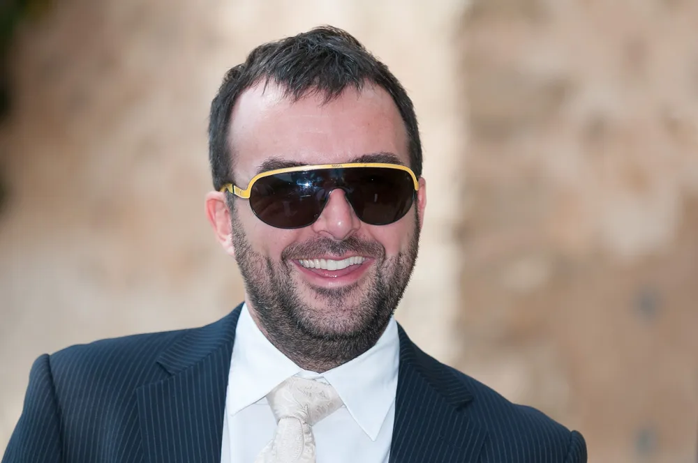 Smiling man with beard wearing gold-rimmed sunglasses, a white dress shirt, paisley tie, and dark pinstripe suit jacket.