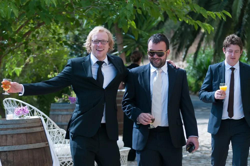 Three men in suits holding drinks outdoors, with the man on the left smiling broadly and spreading his arms.
