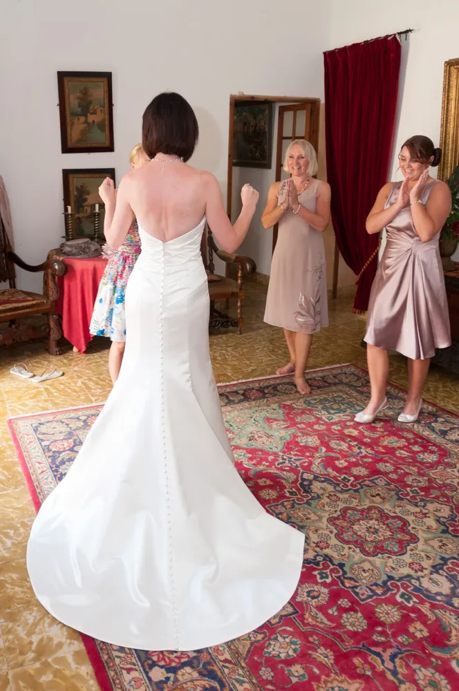 Bride in a white strapless wedding gown seen from the back, with three women in dresses smiling and clapping inside a room with a patterned rug and red curtains.
