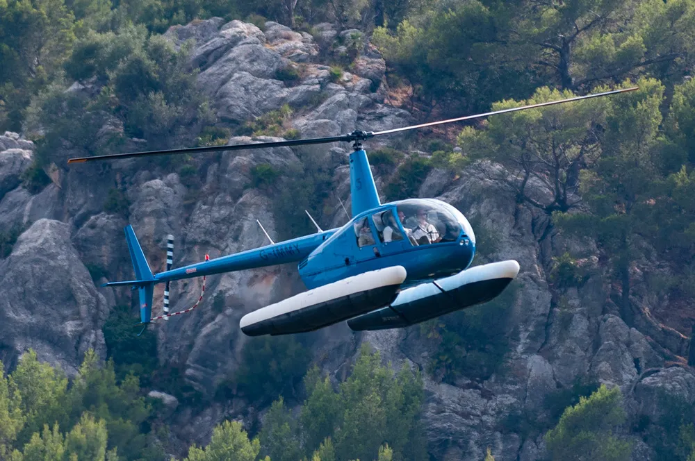 Blue helicopter flying near rocky cliffs with dense trees in the background.