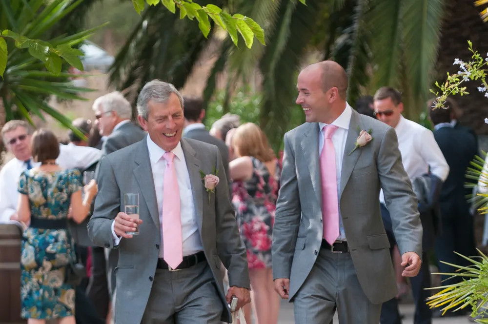 Two men in gray suits with pink ties and rose boutonnieres smiling and walking outdoors at a social event.