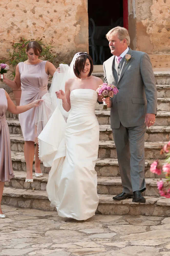 Bride in strapless white wedding gown holding pink and purple bouquet, accompanied by man in gray suit, descending stone steps with bridesmaids in light lavender dresses.