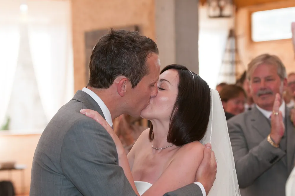 Bride and groom kissing during their wedding ceremony with guests applauding in the background.