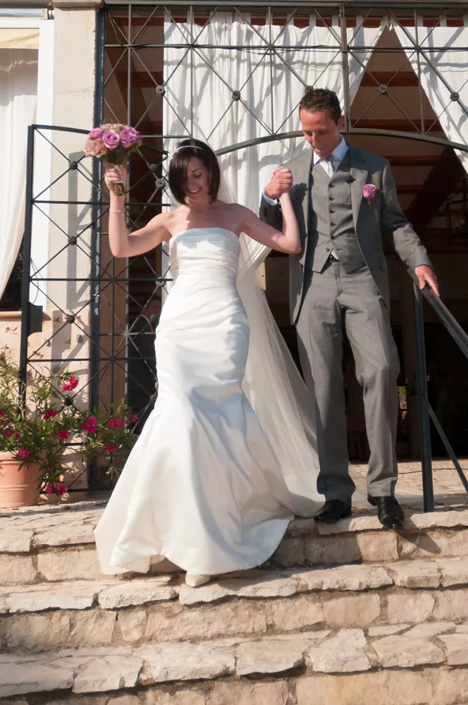 Bride in a white strapless gown holding a bouquet of purple flowers and holding hands with groom in a gray suit as they descend stone steps outside.