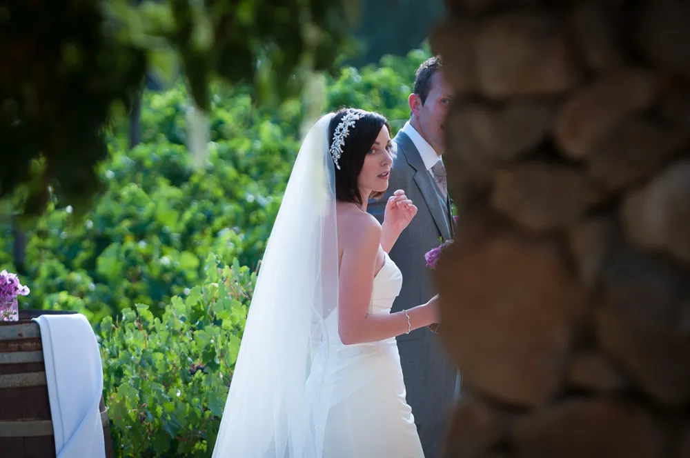 Bride in a white wedding dress and veil standing outdoors with a groom partially visible behind a stone wall.