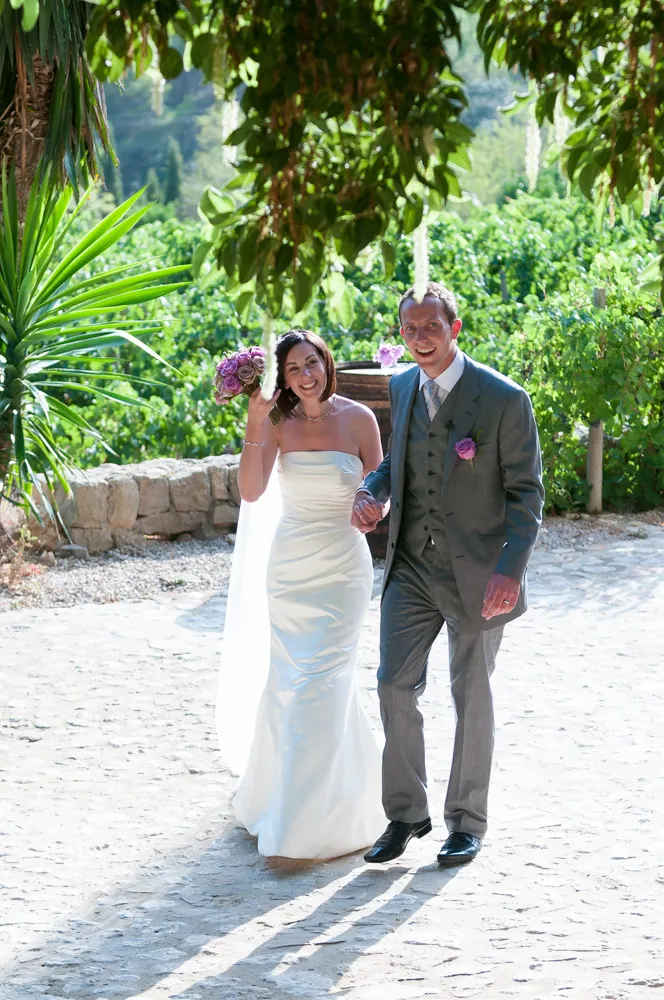 Smiling bride in a white dress holding a purple bouquet and groom in a gray suit walking hand in hand outdoors under leafy trees.