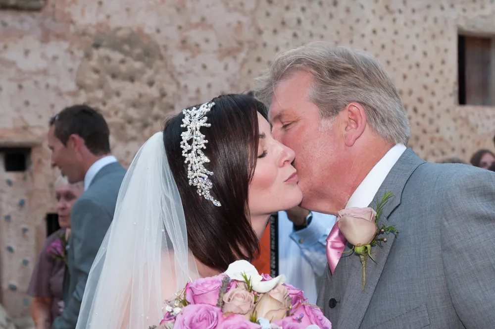 Bride in a white veil and floral headpiece receiving a kiss on the cheek from an older man in a gray suit with a pink tie and rose boutonniere.