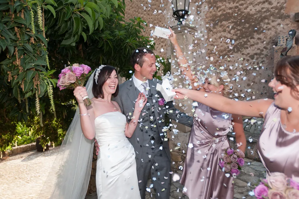 Bride and groom laughing while bridesmaids throw flower petals during outdoor wedding celebration.