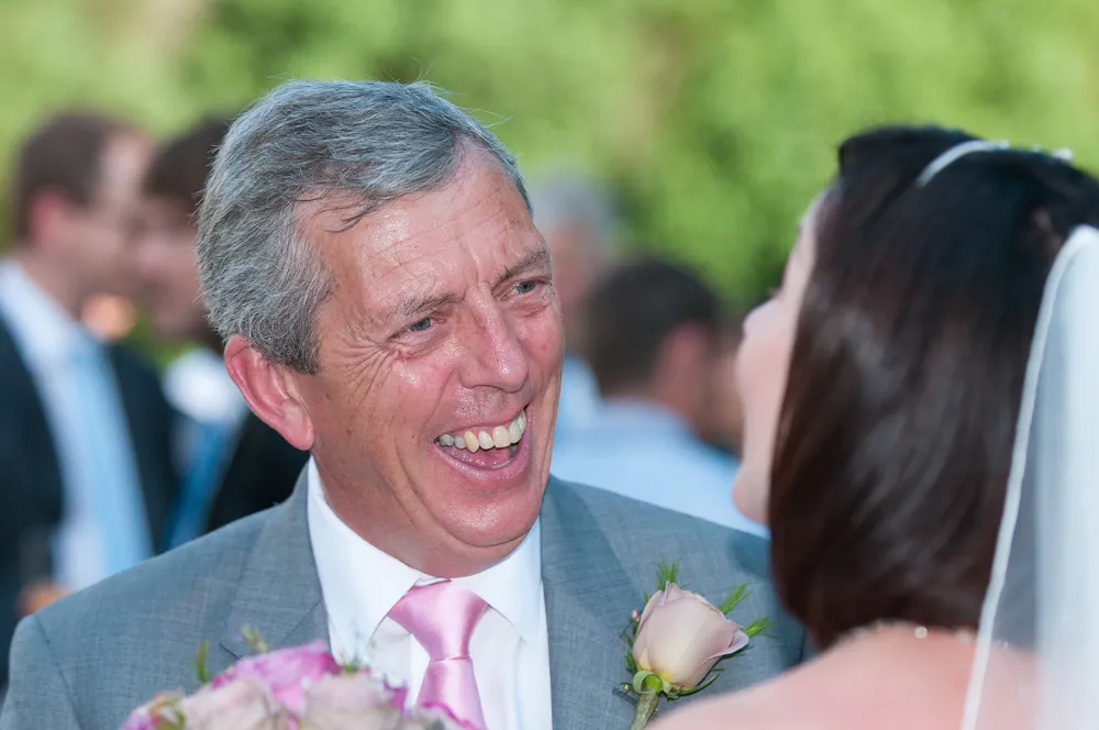 Smiling older man in a gray suit with a pink tie and boutonniere looking at a bride in a wedding veil.