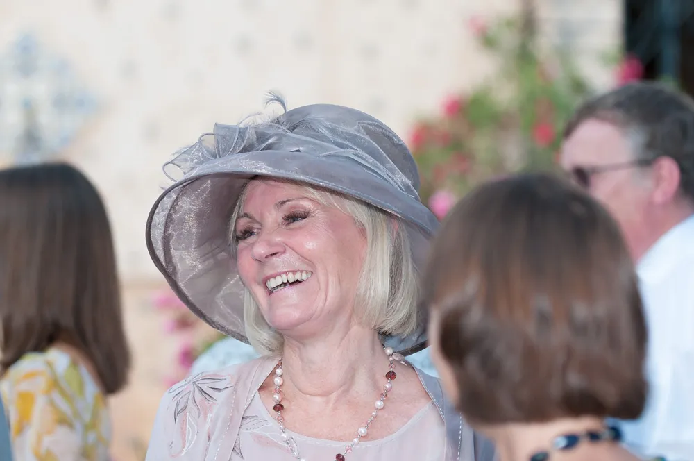 Smiling woman wearing a large silver hat and a necklace, engaged in conversation at a social event.