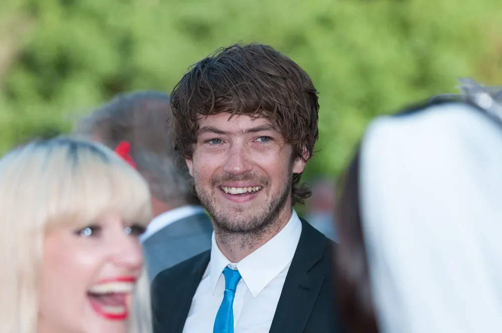Young man with brown hair and beard wearing a suit and blue tie, smiling outdoors among other people.