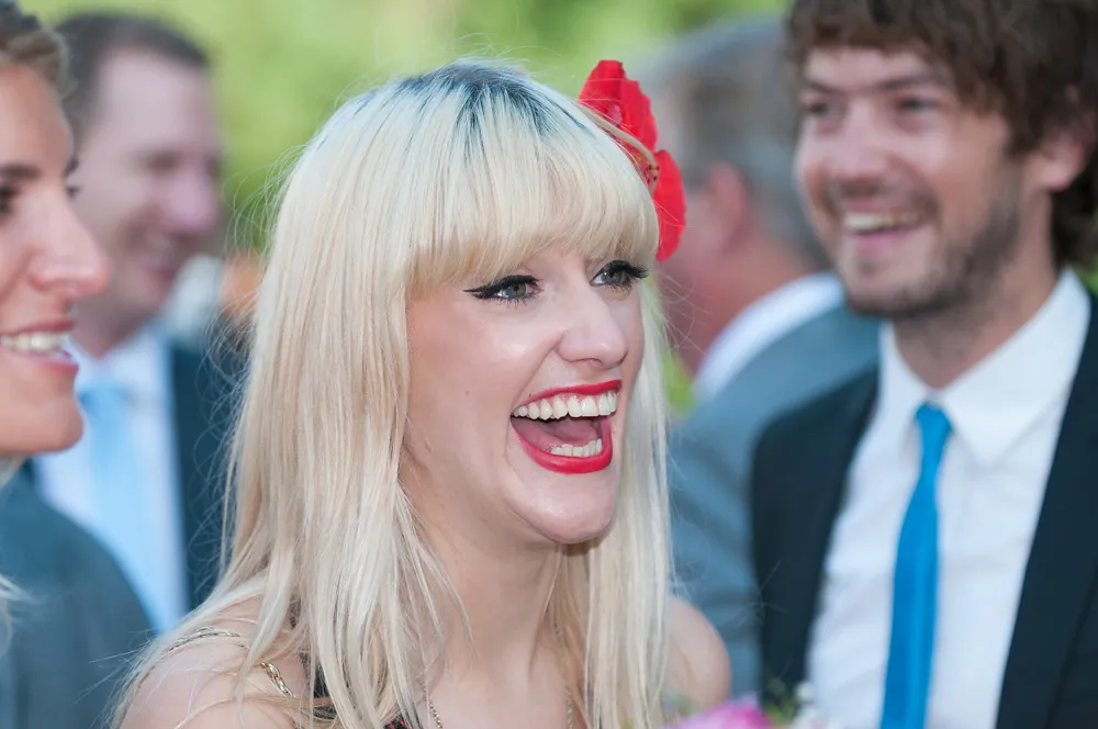Blonde woman with a red flower in her hair laughing among a group of people in formal attire.