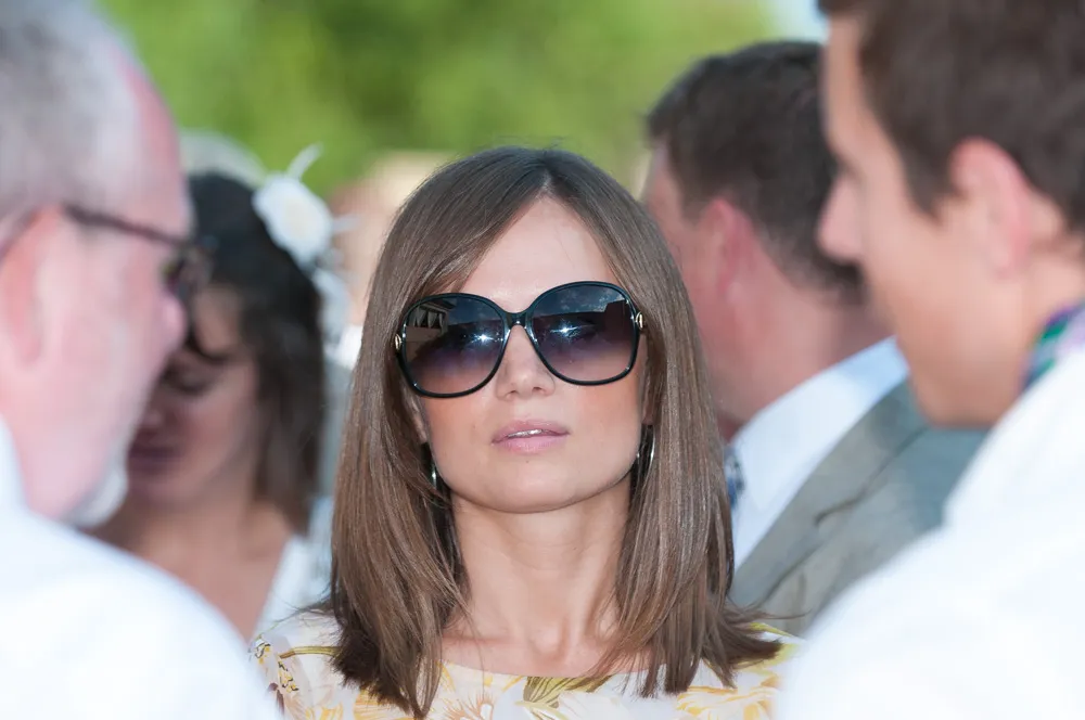Woman with straight brown hair wearing large dark sunglasses and a light floral top at an outdoor event.