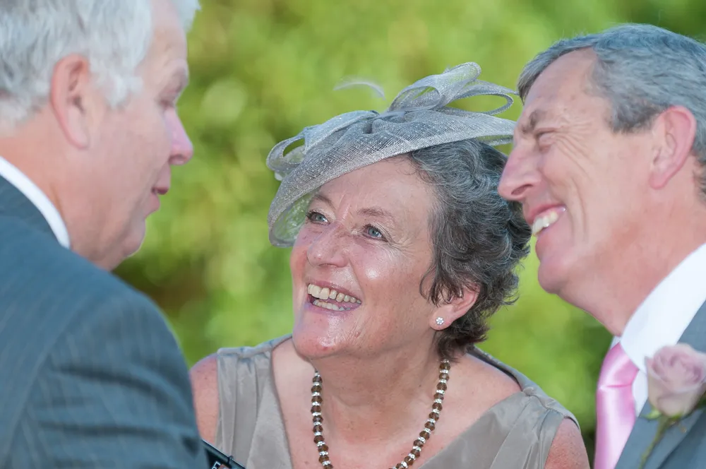 Three elderly people smiling and engaging in conversation outdoors, one woman wearing a decorative gray hat and necklace.