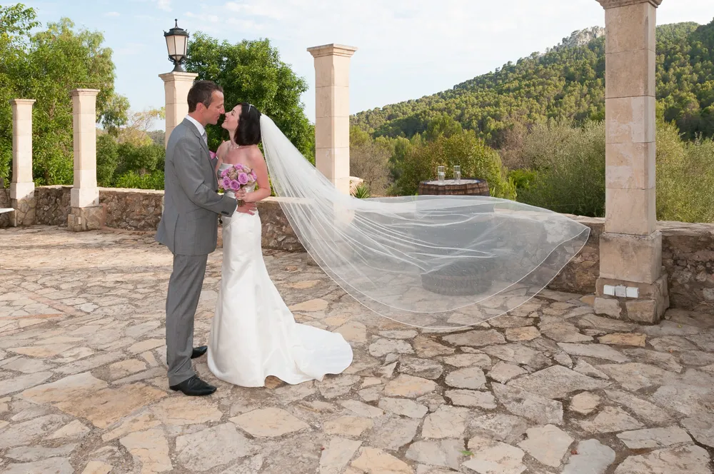 Bride and groom embracing outdoors on a stone patio with the bride's veil flowing behind her and green hills in the background.