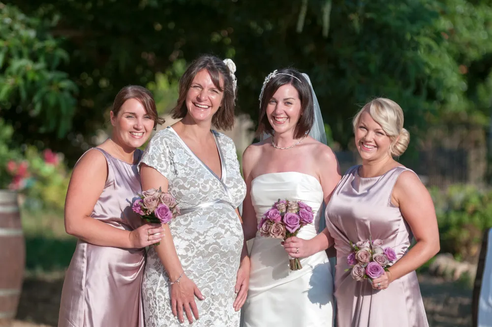 Bride in a strapless white dress holding a bouquet of purple and pink roses, standing with three smiling bridesmaids in light purple satin dresses outdoors.